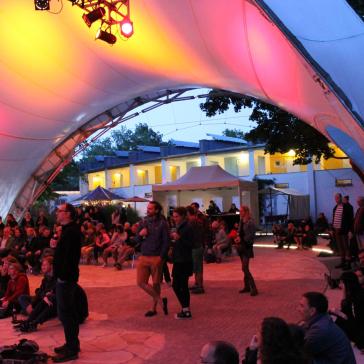 Eine abendliche Veranstaltung unter einem beleuchteten Zeltdach, viele Menschen sitzen und stehen, bunte Lichter, Gebäude im Hintergrund.