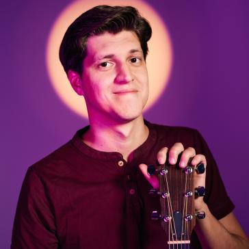  Das Bild zeigt einen jungen Mann, der eine Gitarre hält und sanft lächelt. Er ist in einem lila Licht, das ihn vor einem leuchtenden Kreis hervorhebt.