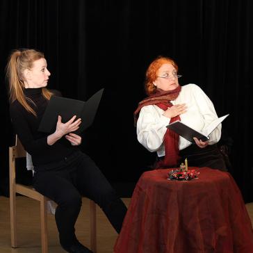  Das Bild zeigt drei Personen in einem Theaterstück. Sie sitzen um einen Tisch, halten schwarze Mappen in den Händen und zeigen lebhafte Gesichtsausdrücke.
