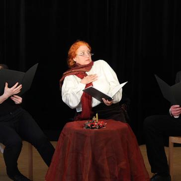  Das Bild zeigt drei Personen in einem Theaterstück. Sie sitzen um einen Tisch, halten schwarze Mappen in den Händen und zeigen lebhafte Gesichtsausdrücke.