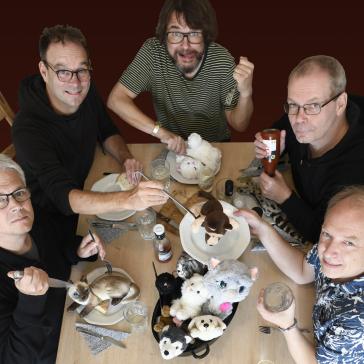  Das Bild zeigt fünf Männer an einem Tisch, die lächelnd in die Kamera blicken. Vor ihnen stehen Teller mit Stofftieren, und einer der Männer hält eine Flasche Ketchup.