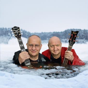 Das zeigt zwei Männer, die in eiskaltem Wasser stehen und Gitarren halten. Der Hintergrund zeigt schneebedeckte Bäume, was auf eine kalte, winterliche Umgebung hinweist.
