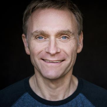  Das Bild zeigt einen lächelnden Mann mit blonden Haaren und blauen Augen vor einem dunklen Hintergrund. Er trägt ein dunkles Oberteil und wirkt freundlich und einladend.