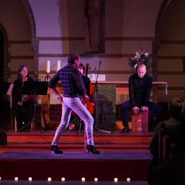 Ein atmosphärisches Bühnenbild in einer Kirche: Ein Mann mit Stock und hohen Absätzen steht im Rampenlicht, begleitet von Musikern und einem Sänger.