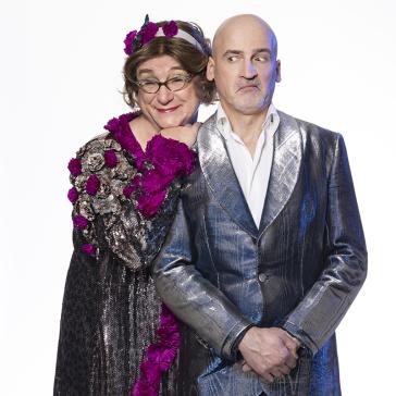  Eine humorvolle Szene: Zwei Personen posieren zusammen. Die eine Person trägt eine auffällige, glitzernde Robe mit violetten Blumen und eine Brille, lächelt liebevoll und legt den Kopf an die Schulter der zweiten Person. Die zweite Person, mit kahlköpfigem Kopf, trägt einen glänzenden silbernen Anzug und schaut skeptisch zur Seite.