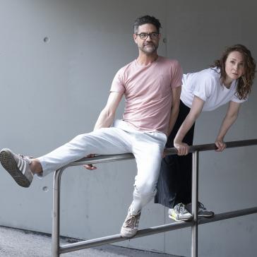Ein Duo in legerer Kleidung posiert spielerisch auf einem Geländer. Eine lockere und kreative Atmosphäre vor einer minimalistischen Betonwand.