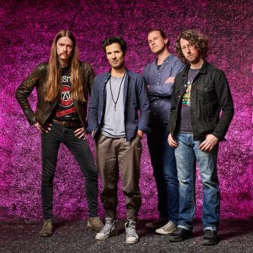 Band-Porträt vor einem leuchtend pinken Hintergrund: vier Mitglieder, lässige Outfits, teils Lederjacke, entspannte Posen mit individueller Ausstrahlung.