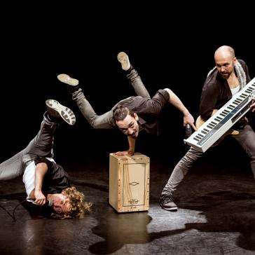 Dynamische Szene einer Performance-Gruppe: ein Akrobat balanciert einhändig, ein Musiker hält ein Keyboard und ein Sänger liegt am Boden, Mikrofon in der Hand.