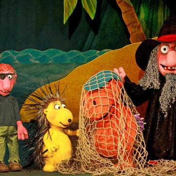 Eine Puppentheater-Szene mit Drachen, Zauberer, Piraten und einem gelben Stacheltier. Farbenfrohes Bühnenbild mit Meer und Insel im Hintergrund.