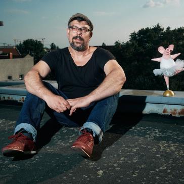 Ein Mann sitzt auf einem Dach und trägt ein schwarzes T-Shirt, Jeans und rote Schuhe. Neben ihm steht eine kleine, rosa Ballerina-Figur.