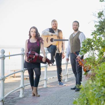 in Trio steht auf einem Gehweg am Meer. Links eine Frau mit einer Drehleier, rechts zwei Männer, einer mit Gitarre und der andere mit Geige.