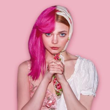 Das Bild zeigt Mädchen, dass links pinke Haare und rechts ein buntes Kopftuch trägt. Sie hat einen ernsten Gesichtsausdruck und hält ihre Hände vor dem Körper zusammen. Der Hintergrund ist in sanften Rosa-Tönen.