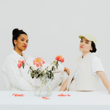 Das Bild zeigt zwei Frauen mit unterschiedlichen Stilen, die entspannt vor einem hellen, neutralen Hintergrund sitzen. Zwischen ihnen steht eine Vase mit Blumen. Beide blicken selbstbewusst in die Kamera.