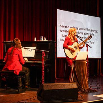 Zwei Musikerinnen führen auf einer Bühne auf. Eine sitzt am Klavier, während die andere Gitarre spielt und singt. Im Hintergrund ist ein großer Bildschirm mit projiziertem Songtext zu sehen. Die Bühne ist mit roten Vorhängen und festlichen Dekorationen geschmückt.