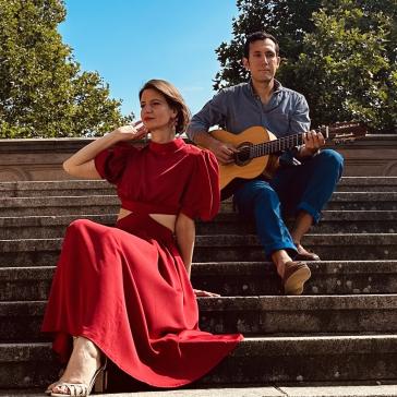 Eine Frau in einem roten Kleid und ein Mann mit einer Gitarre sitzen auf einer Steintreppe im Freien. Der Hintergrund zeigt sonnige Bäume und Architektur.