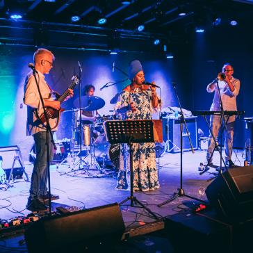 Eine Band spielt auf einer Bühne in einem Club. Links ein Gitarrist, in der Mitte eine Sängerin, rechts ein Trompeter, im Hintergrund ein Schlagzeuger.