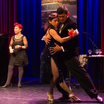 Ein Paar tanzt innig Tango auf einer Bühne, eine Frau in Schwarz singt, und ein Pianist spielt. Bunte Vorhänge im Hintergrund.