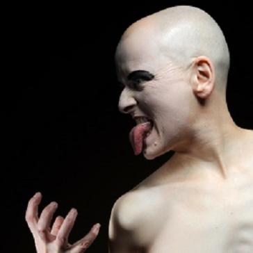 Das Bild zeigt eine Person mit einer glatzköpfigen Frisur und auffälligem Make-up. Sie hat einen provokanten Gesichtsausdruck und streckt die Zunge heraus, während sie mit einer Hand eine ausdrucksstarke Geste macht. Ein rotes Kreuz auf der Brust deutet möglicherweise auf eine künstlerische oder symbolische Bedeutung hin. Der Hintergrund ist schwarz, was die Figur und die Gesichtszüge hervorhebt.