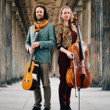 Auf dem Bild sind zwei Musiker zu sehen. Eine Person hält eine Gitarre, die andere ein Cello. Sie stehen in einem überdachten Außenbereich mit mehreren Säulen, was auf einen kulturellen oder historischen Ort hinweisen könnte. Beide tragen stilvolle Kleidung. 