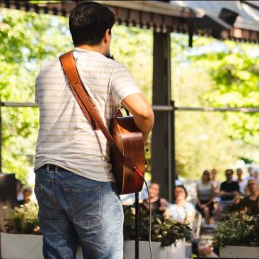 Das Bild zeigt einen Musiker, der mit einer Gitarre auf einer Bühne steht und ein Publikum ansieht. Der Hintergrund ist grün und hell, mit Bäumen und Pflanzen, was eine lebendige Atmosphäre schafft. Das Publikum ist in einer entspannten Position und scheint der Darbietung aufmerksam zuzuhören.