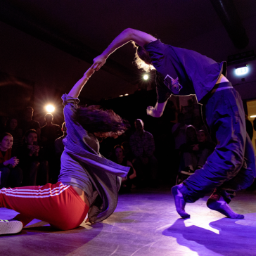 Zwei Tänzer*innen in dynamischer Pose bei einem Battle. Eine Person sitzt auf dem Boden, die andere beugt sich nach hinten. Publikum sitzt im Hintergrund, Bühne in violettem Licht.