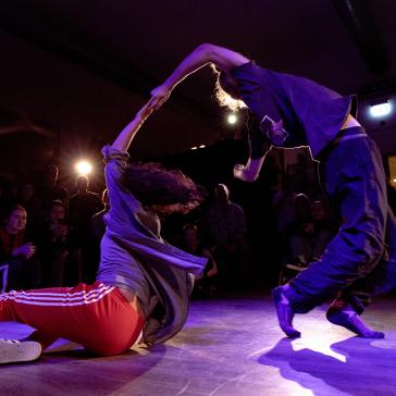 Zwei Tänzer*innen in dynamischer Pose bei einem Battle. Eine Person sitzt auf dem Boden, die andere beugt sich nach hinten. Publikum sitzt im Hintergrund, Bühne in violettem Licht.