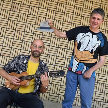 Ein Mann mit Brille spielt Gitarre, während ein anderer daneben steht, eine Mütze hebt und ein T-Shirt mit Donald-Duck-Motiv trägt. Hintergrund mit Muster.