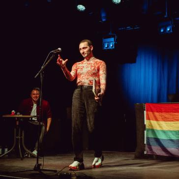 Eine Person in engem, rotem Oberteil und schwarzen Hosen steht auf einer Bühne vor einem Mikrofon, hält ein Buch und gestikuliert. Im Hintergrund sitzt jemand lachend an einem Tisch. Regenbogenflagge seitlich.