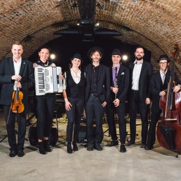 Eine Band aus sieben Menschen in eleganter Kleidung steht in einem Backsteingewölbe. Sie halten Instrumente wie Geige, Akkordeon, Klarinette und Kontrabass und lächeln in die Kamera.