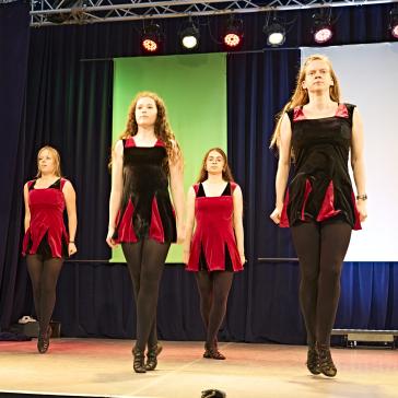 Das Bild zeigt eine Gruppe von Tänzerinnen, die auf einer Bühne auftreten. Sie tragen schwarz-rote Kleider und schwarze Strumpfhosen. Der Hintergrund ist blau mit einem grünen Akzent, und bunte Beleuchtung schafft eine lebhafte Atmosphäre. Die Szene deutet auf eine traditionelle Tanzvorführung hin.