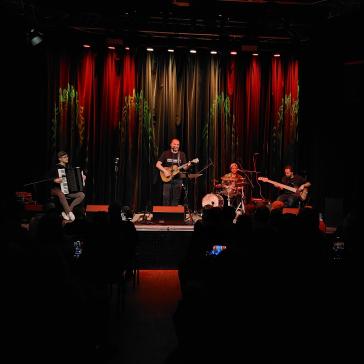 Eine vierköpfige Band spielt auf einer Bühne mit roter und grüner Beleuchtung. Ein Musiker steht mit Gitarre, während Akkordeon, Schlagzeug und Bass gespielt werden. Das Publikum sitzt im Dunkeln.