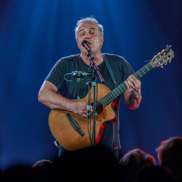 Ein Musiker mit grauen Haaren und schwarzem Shirt spielt auf einer Akustikgitarre und singt in ein Mikrofon. Blaue Bühnenlichter strahlen ihn an, während das Publikum als dunkle Silhouetten zu sehen ist.