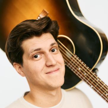 Ein junger Mann mit hellbeigem T-Shirt schaut lächelnd in die Kamera. Er trägt eine akustische Gitarre über der Schulter. Der Hintergrund ist neutral und hell.