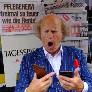 Ein Mann im blauen Nadelstreifenanzug sitzt vor Zeitungen, hält zwei Handys in den Händen und schaut mit weit geöffnetem Mund überrascht oder schockiert auf die Bildschirme.