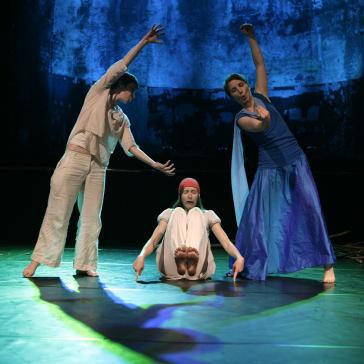 Das Bild zeigt drei Tänzer auf einer Bühne, die eine ausdrucksstarke Choreografie aufführen. Zwei der Tänzer sind stehend und bewegen sich anmutig, während der dritte auf dem Boden sitzt. Die Kostüme sind in Weiß- und Blautönen gehalten. Der Hintergrund ist dunkel, mit blauen Akzenten, die eine künstlerische und eindrucksvolle Atmosphäre schaffen.