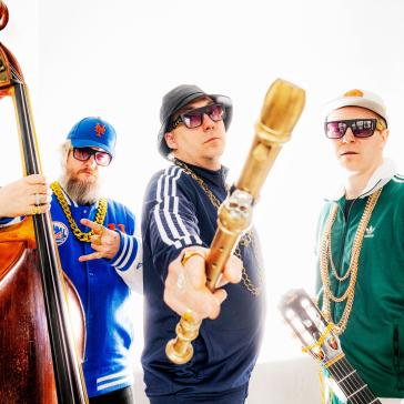 Drei Männer in Sportkleidung und Goldketten posieren cool mit Musikinstrumenten, darunter ein Kontrabass und eine Gitarre. Einer hält eine große Blockflöte in die Kamera.