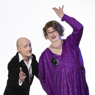 Zwei Personen posieren fröhlich: links ein Mann in schwarzem Glitzeranzug, rechts eine Person in lila Glitzerkleid mit Perlenkette, Brille und erhobener Hand in verspielter Pose.