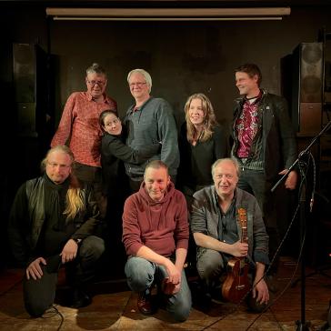 Acht Menschen posieren fröhlich auf einer kleinen Bühne für ein Gruppenfoto. Drei Personen knien vorn, einer hält eine Ukulele, fünf stehen dahinter. Die Stimmung ist warm und freundschaftlich.