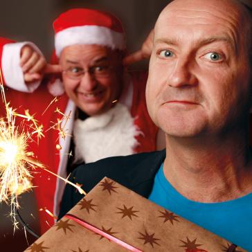 Ein Mann mit ernstem Blick hält ein Geschenk, im Hintergrund hält ein fröhlicher Mann im Weihnachtsmannkostüm die Ohren zu, während eine Wunderkerze hell aufleuchtet.