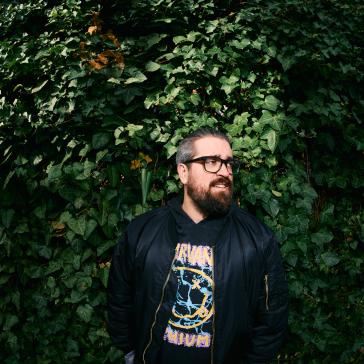 Ein Mann mit Bart und Brille steht vor einer dichten grünen Efeuhecke. Er trägt eine schwarze Jacke und ein Nirvana-Shirt und blickt lächelnd zur Seite.