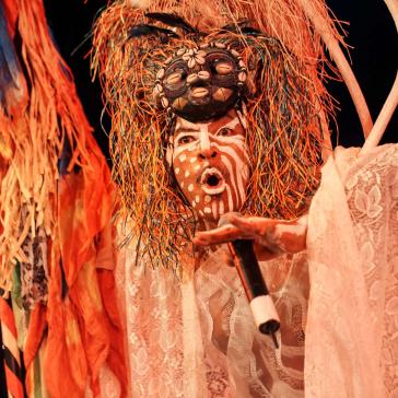 Person mit bemaltem Gesicht, bunter Masken-Kopfbedeckung und Spitzengewand singt oder spricht expressiv ins Mikrofon bei einer Bühnenperformance mit farbenfrohem Dekor.