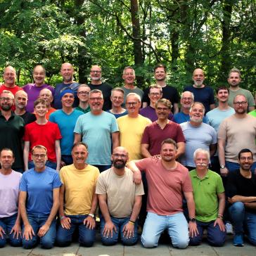Eine große Gruppe von Männern in bunten T-Shirts steht und kniet lachend im Grünen für ein Gruppenfoto im Wald.