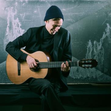 Ein junger Mann mit dunklem Anzug und Mütze sitzt lächelnd auf einer Bank und spielt Gitarre vor einer rauen, industriellen Wand.