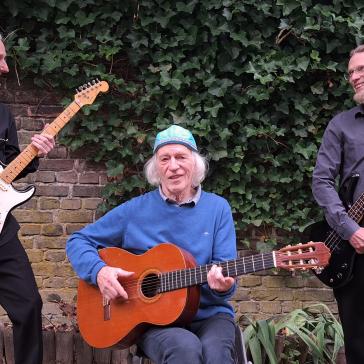 Drei ältere Männer stehen vor einer efeubewachsenen Mauer. Zwei stehen mit E-Gitarren, einer sitzt mit Akustikgitarre. Alle lächeln, die Stimmung ist entspannt und musikalisch.