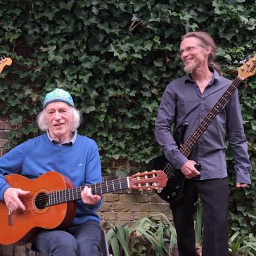 Drei ältere Männer stehen vor einer efeubewachsenen Mauer. Zwei stehen mit E-Gitarren, einer sitzt mit Akustikgitarre. Alle lächeln, die Stimmung ist entspannt und musikalisch.