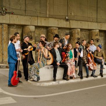 Eine Gruppe von Musikern sitzt und steht auf einem Bordstein vor einer Betonwand, spielt verschiedene Instrumente und lacht zusammen. Es wirkt wie ein fröhliches Straßenkonzert.