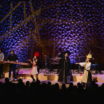Achtköpfige Band auf Bühne mit traditionell-kreativen Outfits, bunter Beleuchtung und koreanischen Instrumenten, singend und musizierend vor Publikum.