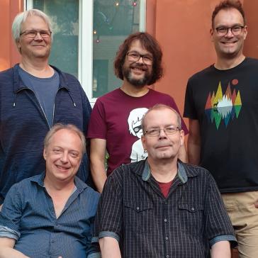 Fünf Männer stehen und sitzen lächelnd in einem Garten vor einer orangefarbenen Wand mit Fenster. Es ist ein Gruppenfoto in entspannter, freundlicher Atmosphäre.