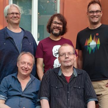 Fünf Männer stehen und sitzen lächelnd in einem Garten vor einer orangefarbenen Wand mit Fenster. Es ist ein Gruppenfoto in entspannter, freundlicher Atmosphäre.