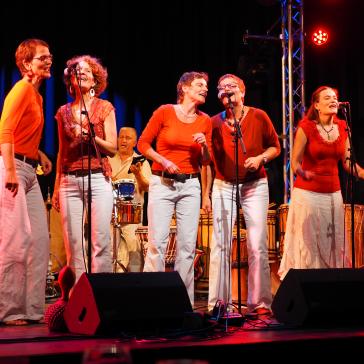 echs Personen in roten Oberteilen und weißen Hosen singen fröhlich auf einer Bühne. Im Hintergrund spielen Musiker Percussion-Instrumente. Bunte Lichter beleuchten die Szene.